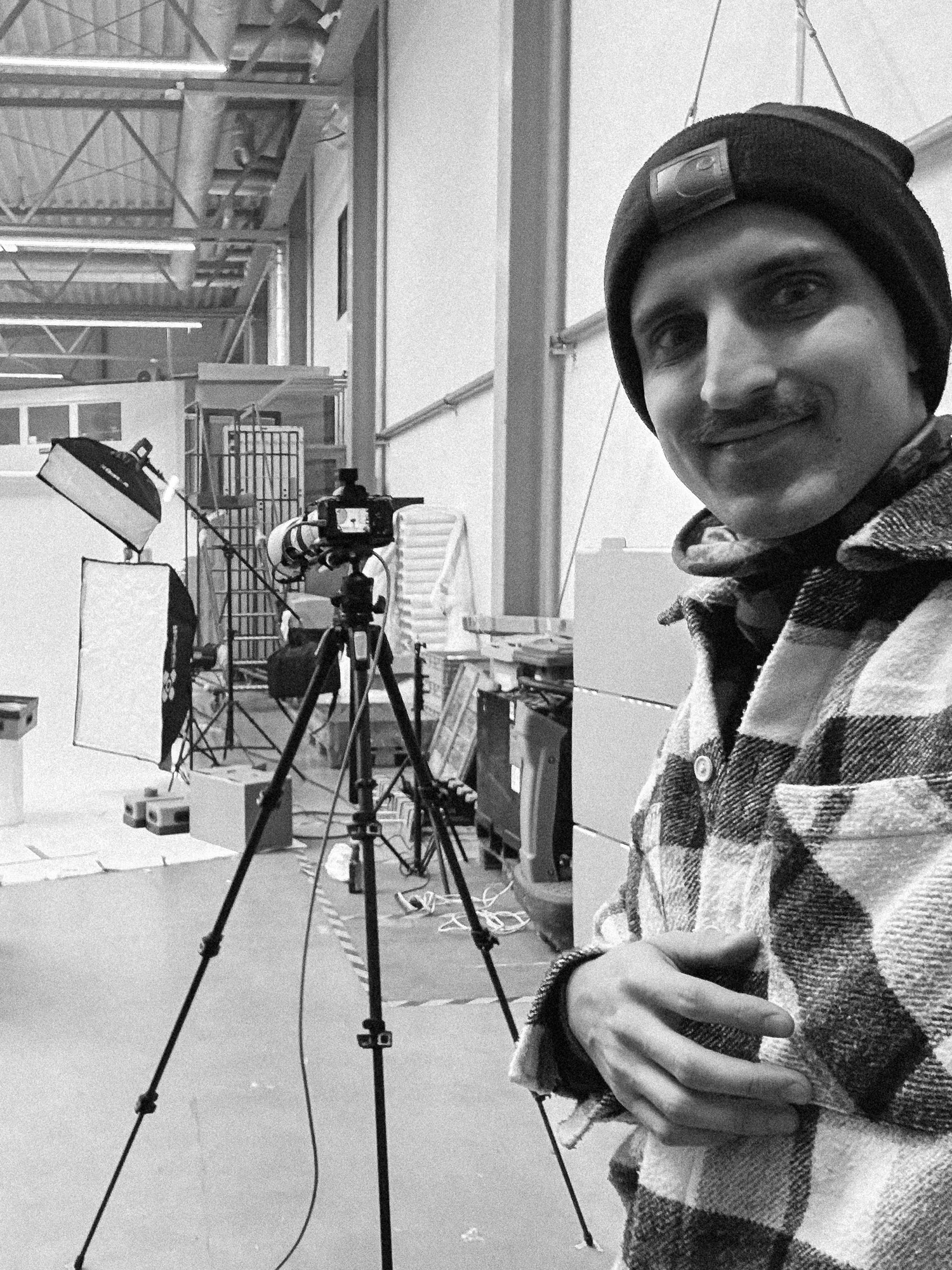 Adam Ziarko na planie sesji DANPLA — selfie z uśmiechem, laptop tether i mobilne studio w hali, kadr B&amp;W