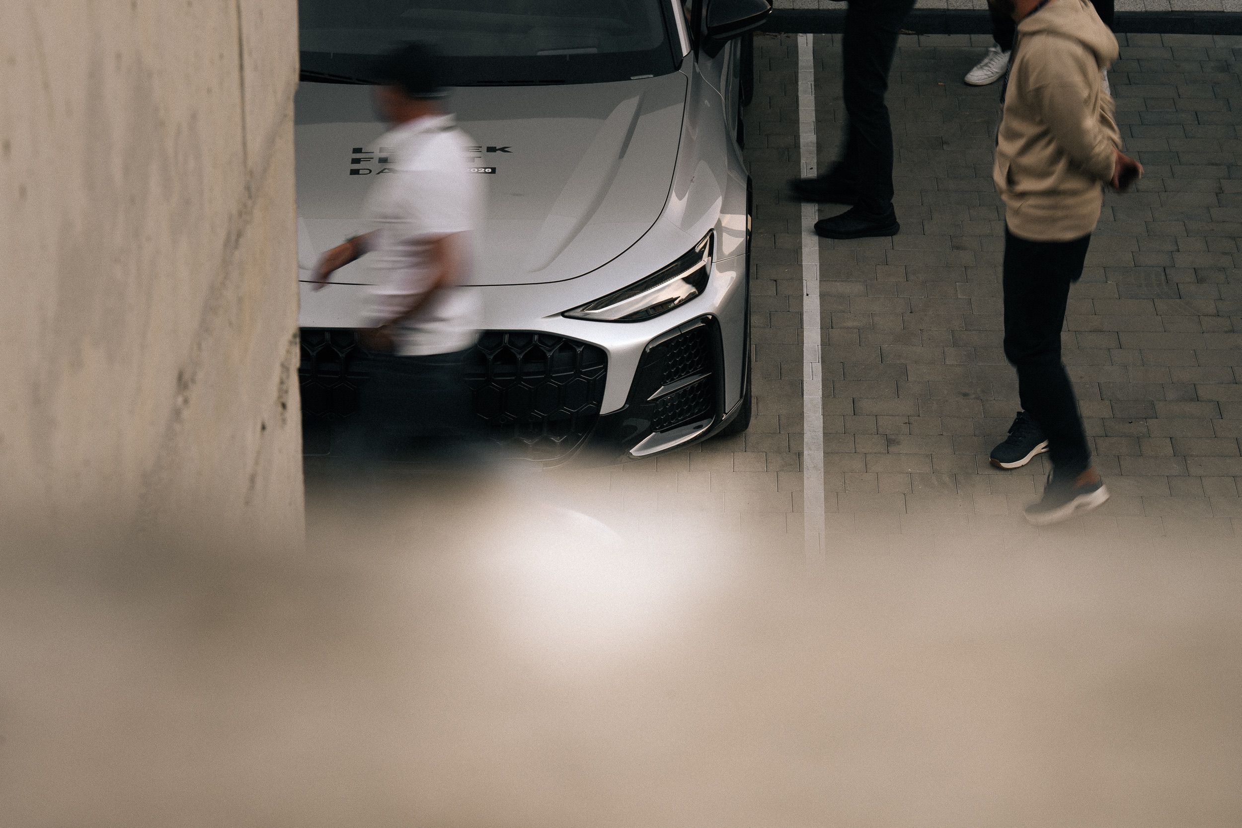 Aufnahme hinter einer Wand hervor: Gäste um einen silbernen Audi auf dem Parkplatz