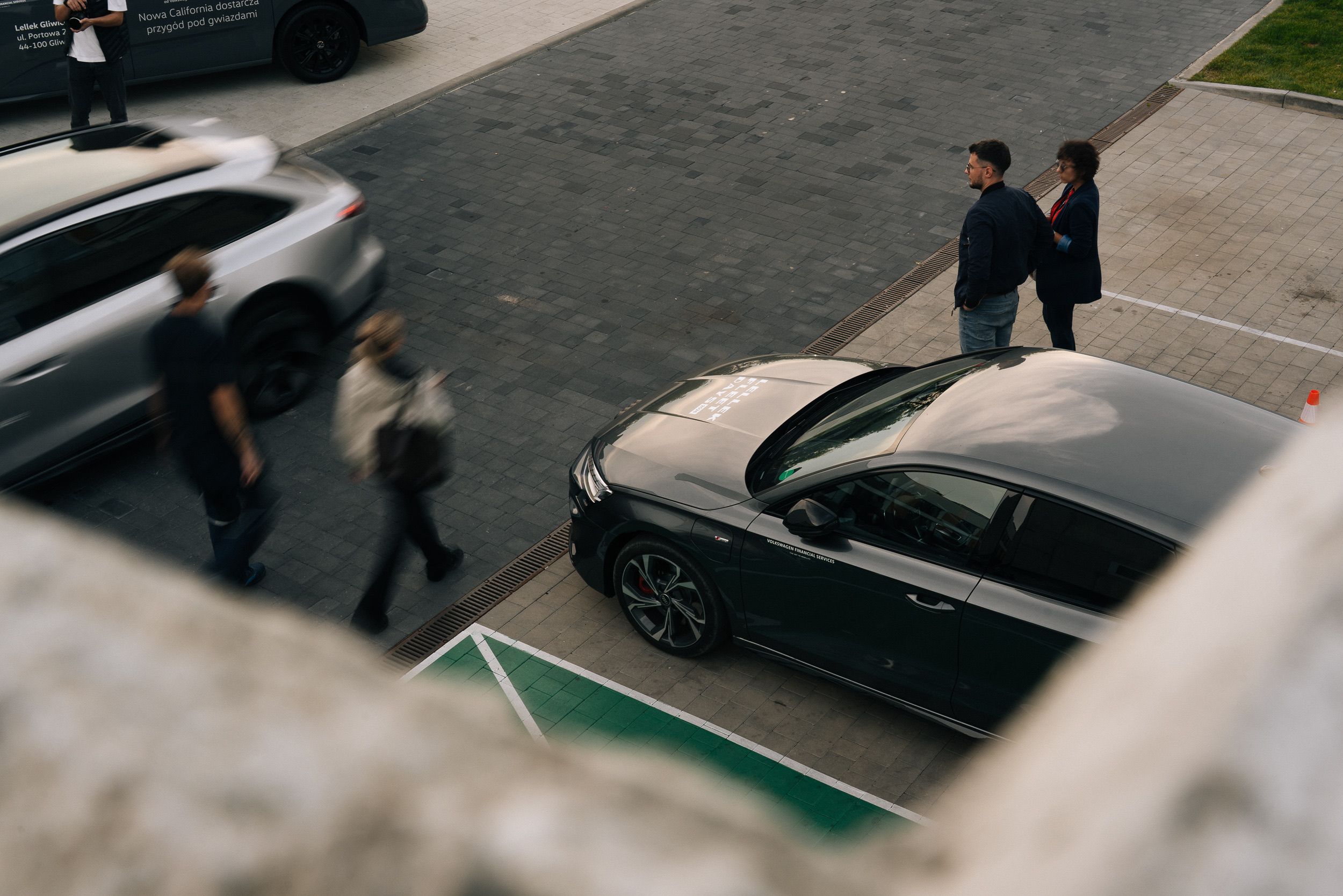 Blick von oben über eine Betonmauer: Teilnehmer gehen und unterhalten sich zwischen den Autos auf dem Parkplatz
