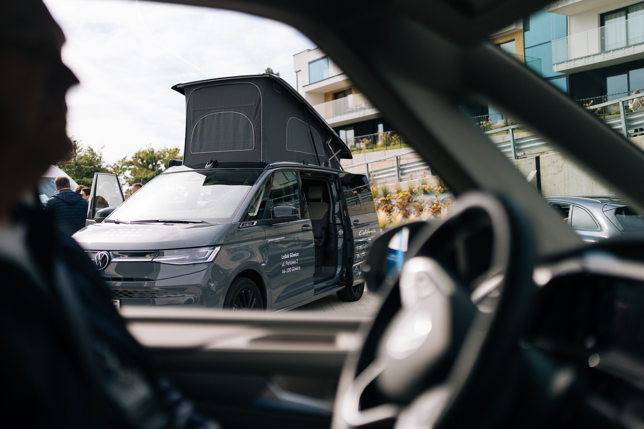 Aus der Kabine: Silhouette des Fahrers und Lenkrad, hinter der Scheibe ein VW California mit aufgestelltem Dach und offenen Türen vor dem Hotel