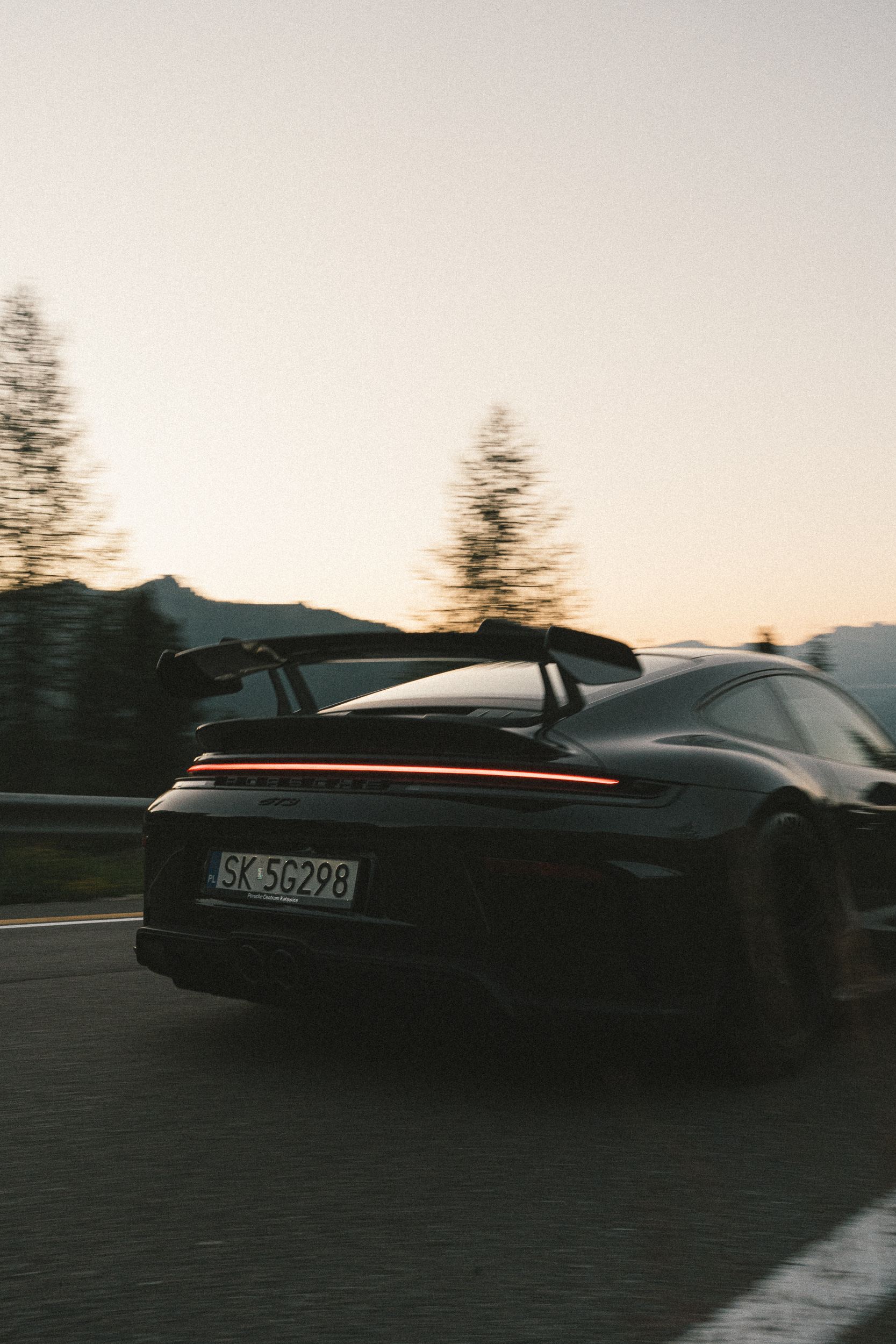 Tył 911 GT3 z zapaloną listwą świateł na górskiej drodze, za nim świerki i jasne niebo nad Dolomitami — kadr pionowy