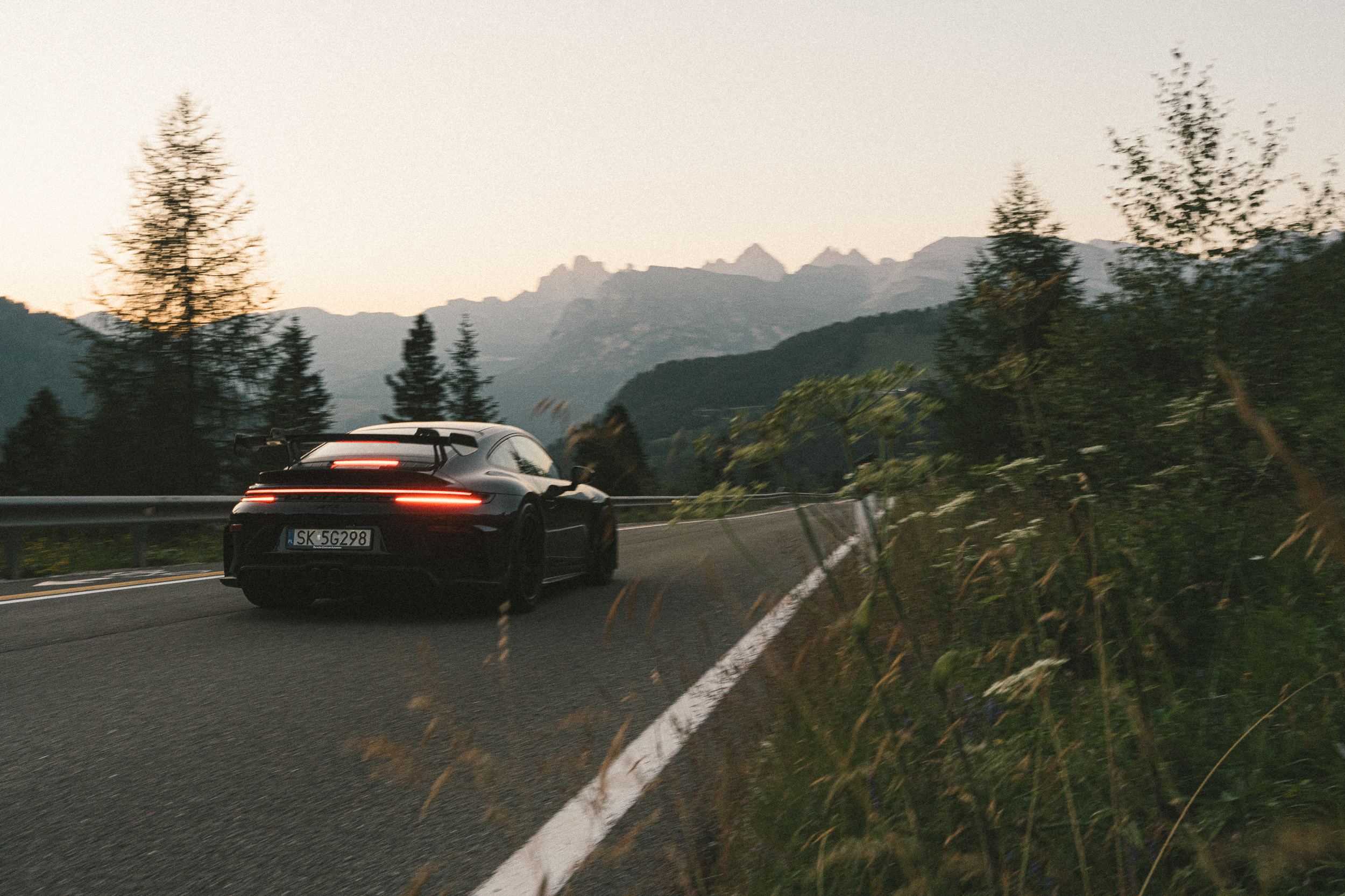 911 GT3 z zapalonymi światłami odjeżdża w zakręt między trawami, w tle Dolomity pod jasnym niebem