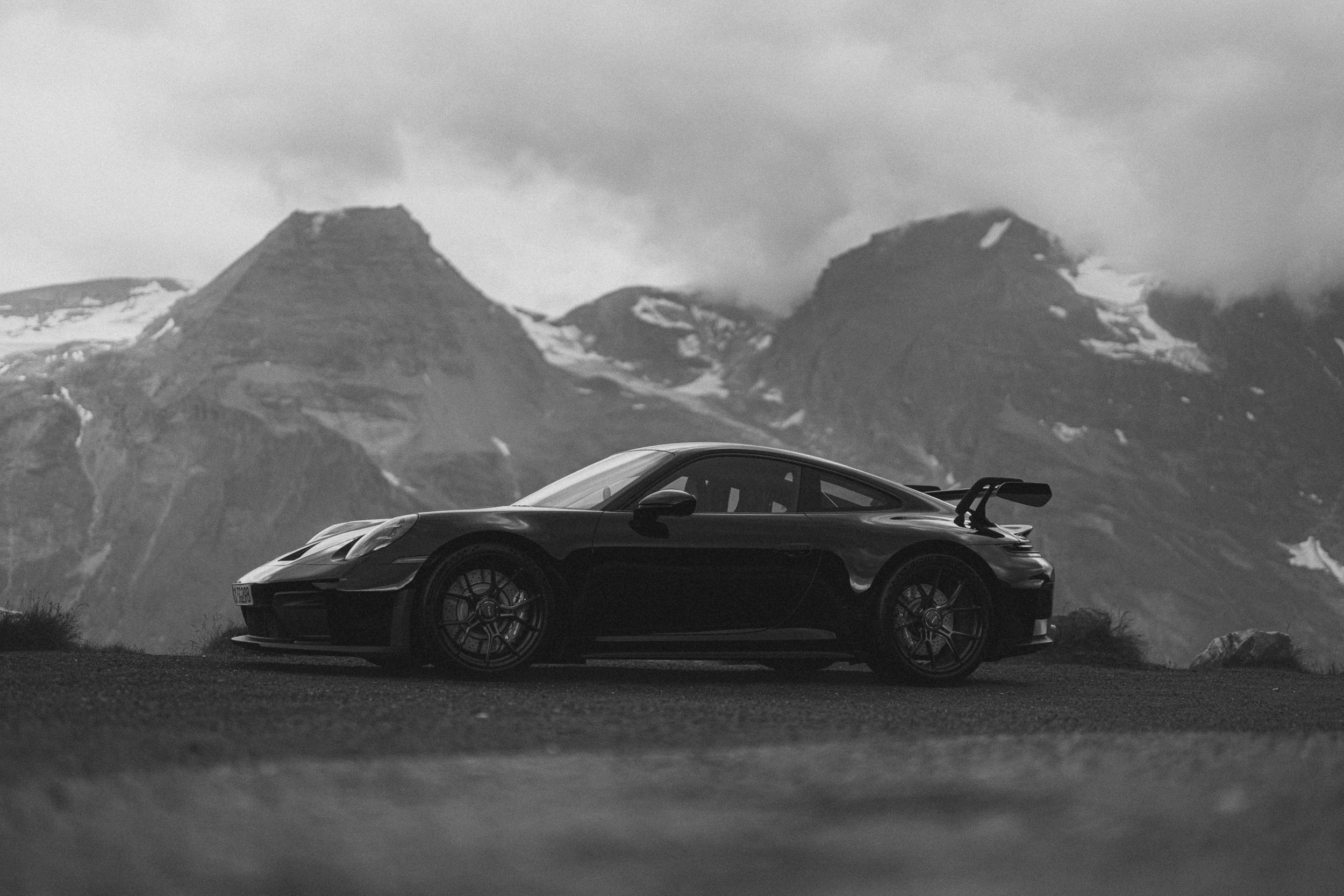 Czarne Porsche 911 GT3 bokiem na tle ośnieżonych szczytów Großglocknera w chmurach — kadr czarno-biały