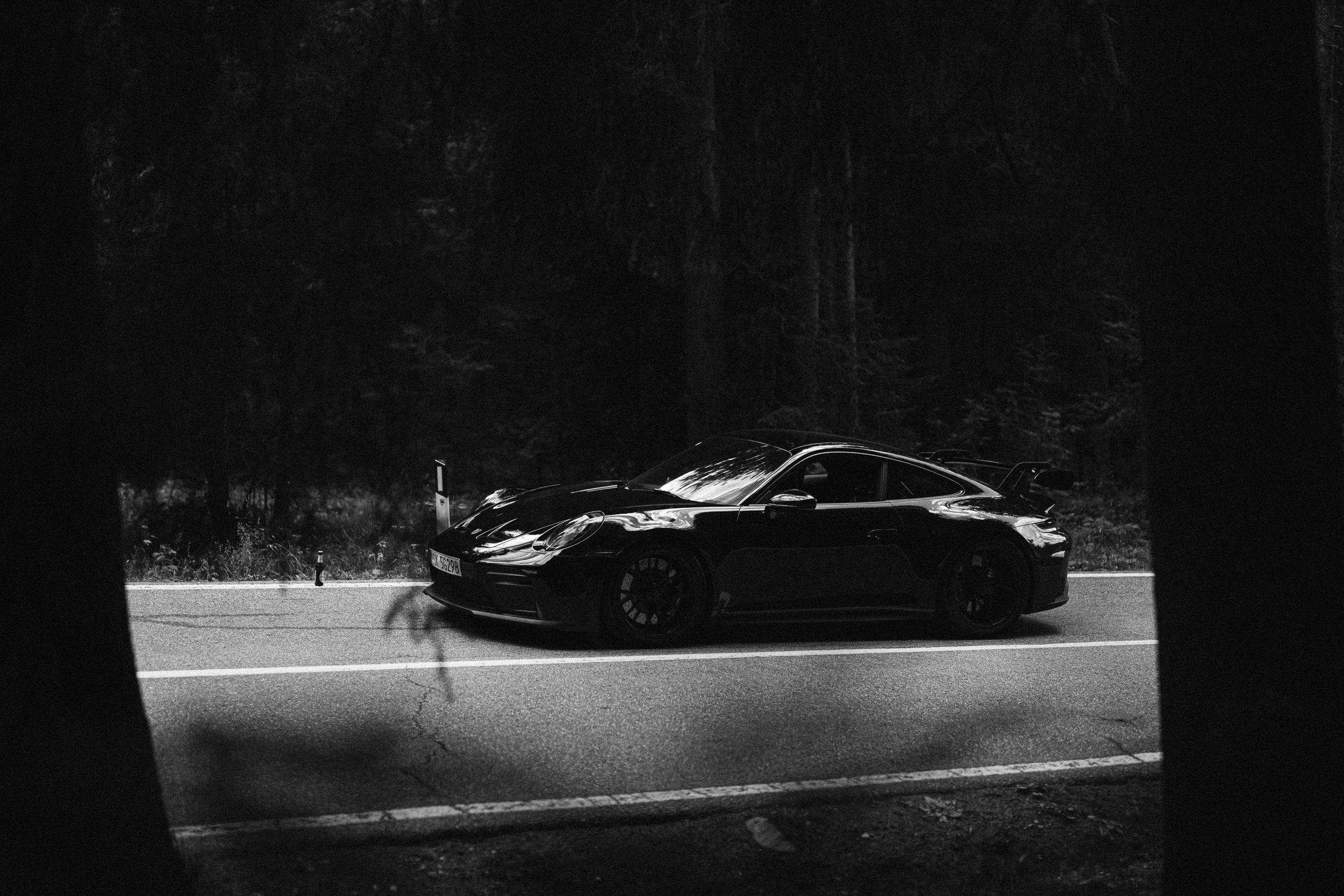 911 GT3 bokiem w ostrym świetle na drodze, ujęte w prześwicie między ciemnymi pniami drzew — kadr czarno-biały