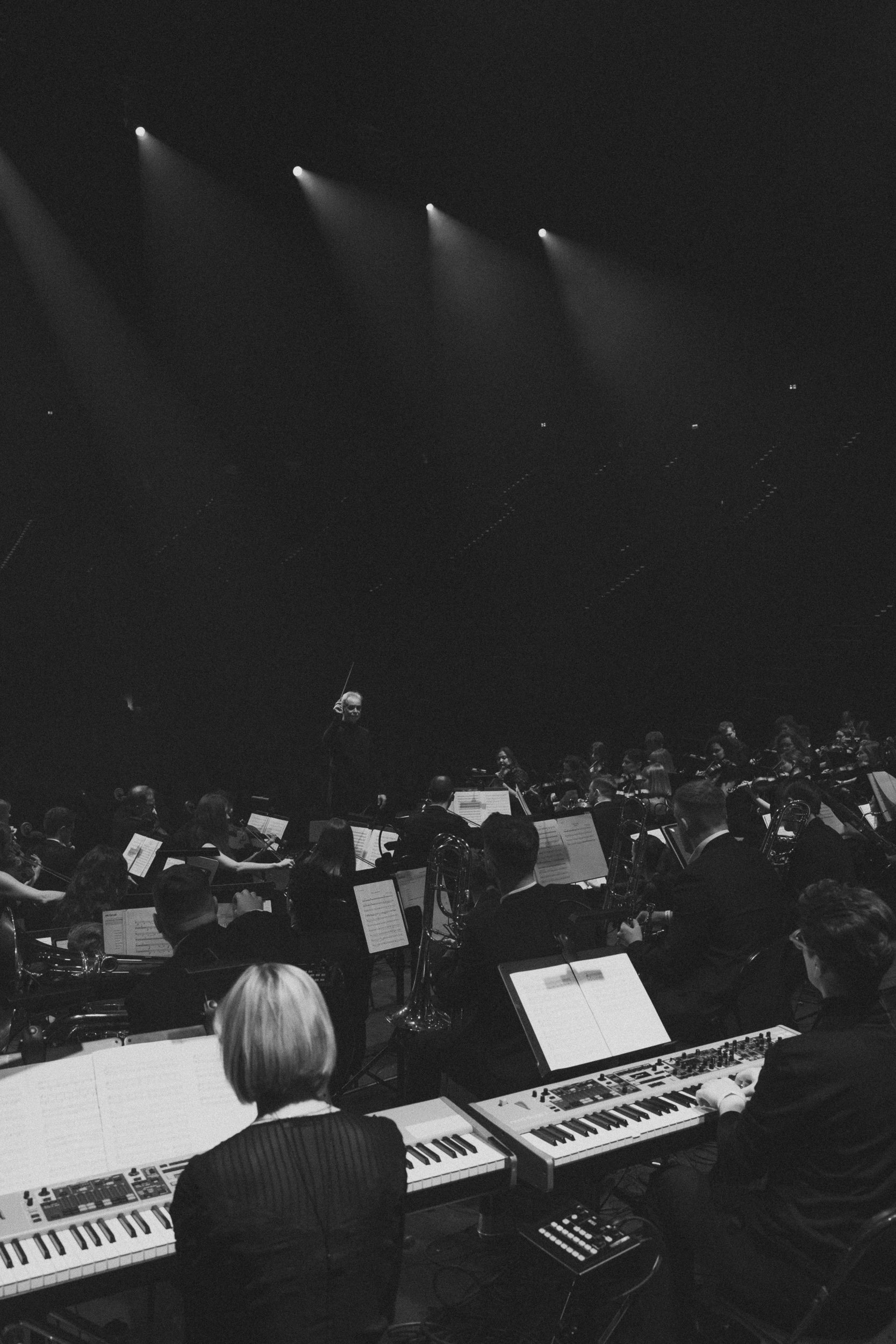 Klawiszowcy przy instrumentach na próbie, w głębi orkiestra pod reflektorami — kadr czarno-biały