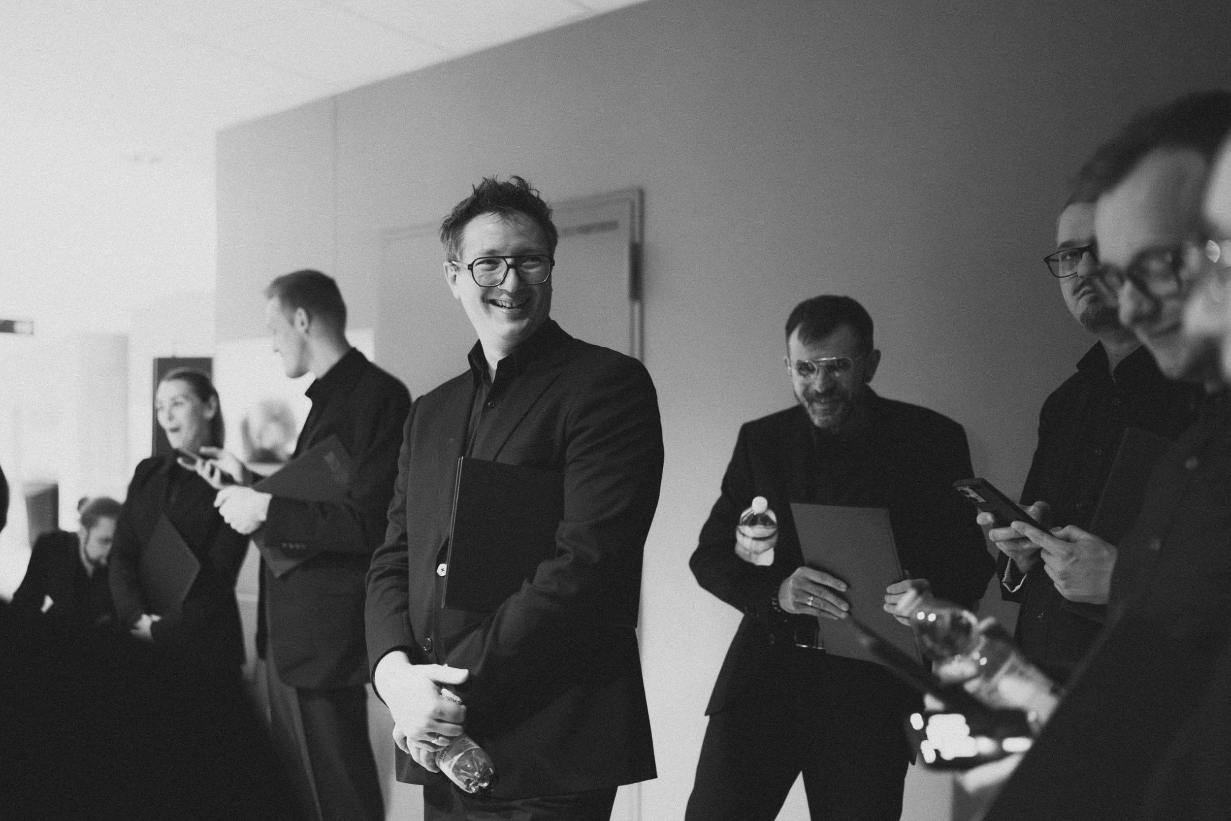 Muzycy w czarnych marynarkach śmieją się w korytarzu areny — kadr czarno-biały