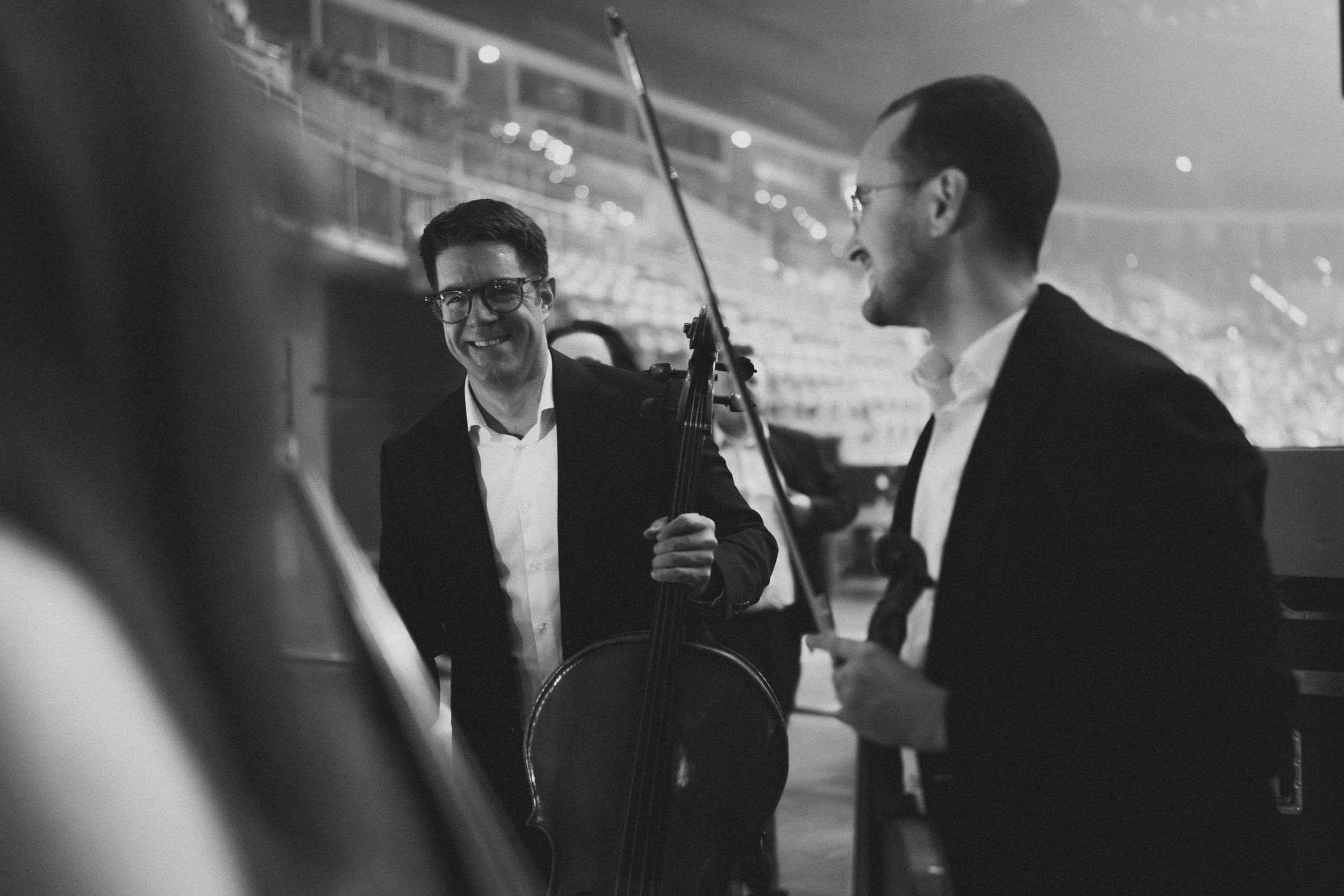 Dwóch muzyków w czarnych marynarkach śmieje się za sceną z instrumentami w dłoniach, w tle trybuny areny — kadr czarno-biały